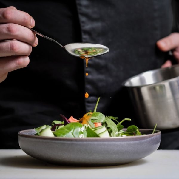 Close-up of the hands of a male chef on a black background. Pour sauce from the spoon on the salad dish. Food decoration.